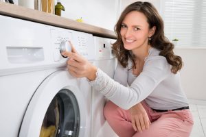 תמונת נושא של המאמר על תיקון מכונת כביסה בו רואים אישה מחייכת מול מכונת כביסה ולוחצת על כפתור התוכניות