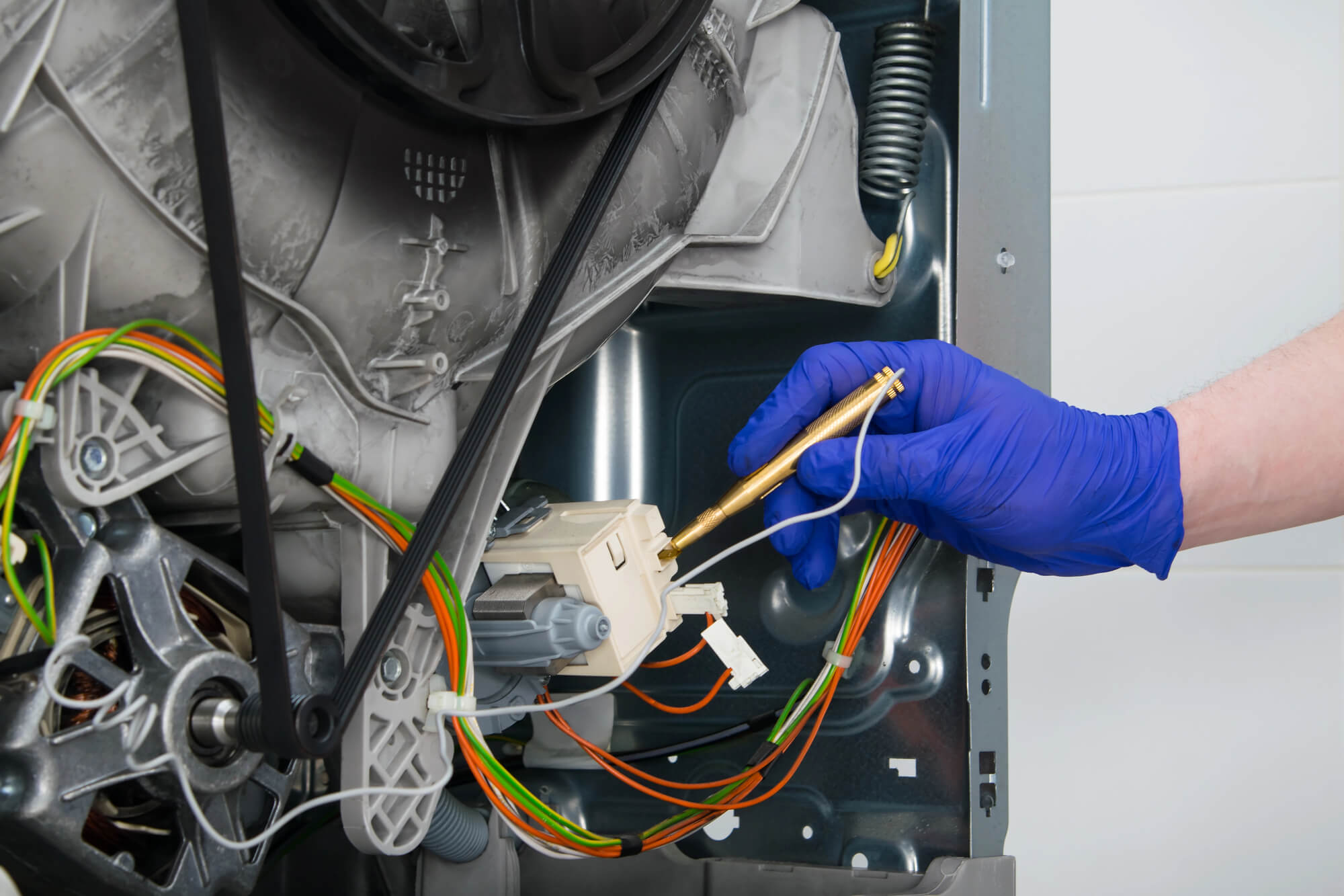 תמונת נושא של המאמר על טכנאי מכשירי חשמל ביתיים המציגה יד עם כפפה כחולה בודקת או מתקנת את מנוע ורכיבים פנימיים של מכונת כביסה עם חגורה כבלים סלילים וקפיץ