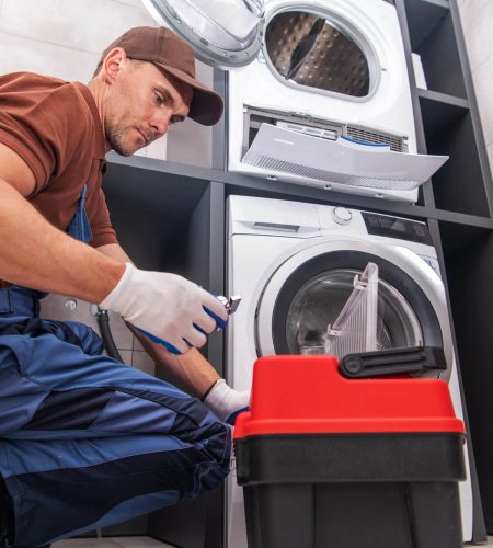 תמונת נושא שנייה של דף השירות טכנאי מייבשי כביסה המציגה טכנאי מכשירי חשמל לובש כפפות כובע ואוברול כורע מול מכונת כביסה בחדר אמבטיה עם אריחים הוא מתקן אותה ליד מייבש פתוח.